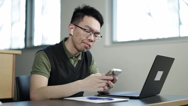 A young Asian male student wearing glasses and earbuds puts down his studies to scroll on his smartphone while sitting at a desk with a laptop, ideal for student distraction, social media addiction, o