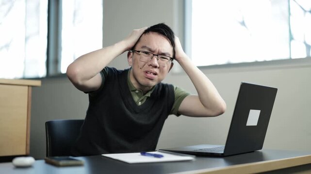 A young Asian male student wearing glasses grabs his head in shock and frustration while looking at his laptop, ideal for student stress, bad news, or academic setback concepts.