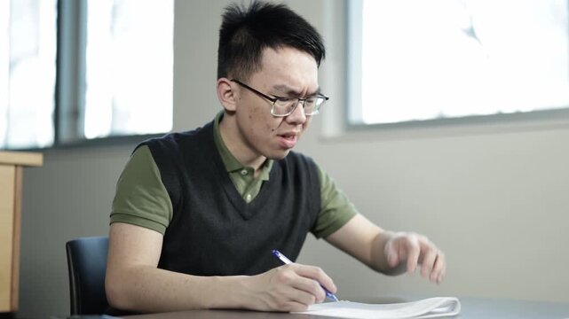 A young Asian male student wearing glasses scratches his head with a confused expression while attempting a test or exam at a desk, ideal for student confusion, exam stress, or academic challenge.