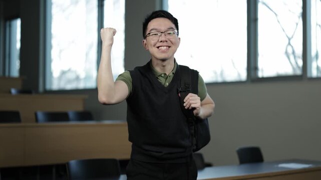 A young Asian male student wearing glasses and a backpack pumps his fist in celebration in an empty lecture hall, ideal for student success, academic achievement, or personal victory concepts.