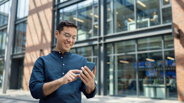 Happy young Hispanic business man manager using financial banking mobile apps on cell phone standing in city, professional businessman holding smartphone in hands looking at cellphone outdoor office.