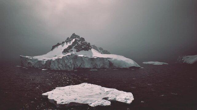 Lone iceberg and nearby floating ice on dark cold water, jagged snowcapped ridges under overcast sky, mood reflective and austere, emphasizes environmental