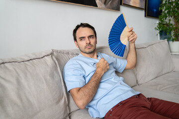 Sad man fanning himself with blue fan in apartment. Exhausted guy suffering from heat, sitting on couch, no air conditioner at home. Hot temperature, bad summer weather, climate control concept