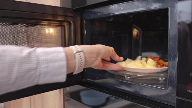 Woman holds plate of food and places it into built in microwave oven in kitchen. Concept of reheating meal and quick home cooking
