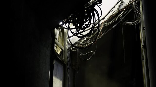 Tangled cables above broken window, dim light slicing through grime, frayed insulation draped over warped frame, cracked plaster and dustcoated surfaces,