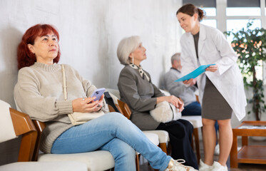 Quiet senior woman waiting for medical appointment as nurse provides assistance to group in corridor