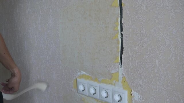 Hand removes wallpaper from wall, revealing electrical outlets and surface damage