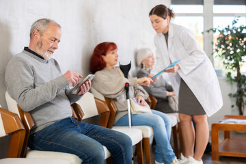 Obraz premium Calm elderly man sitting in clinic queue while nurse talks to other patients in background
