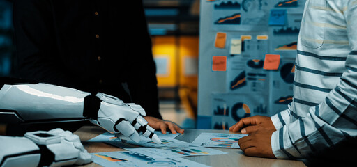 Naklejka na ściany i meble A human and a robotic arm engage in a collaborative discussion in a modern office, analyzing data charts and visuals to enhance productivity and innovation.Noogenesis