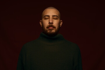 Obraz premium Close-up portrait of a calm bald man with a beard and closed eyes, wearing a dark green turtleneck sweater, posing against a deep burgundy background with dramatic, moody studio lighting.