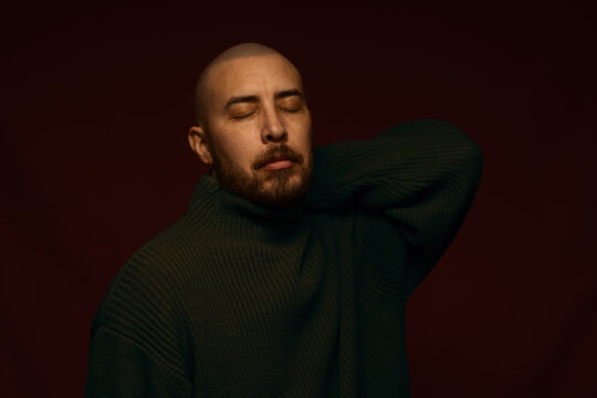 Bald man with beard wearing a dark olive green turtleneck sweater, eyes closed in a calm, contemplative pose against a moody dark red background, studio portrait with dramatic low key lighting.