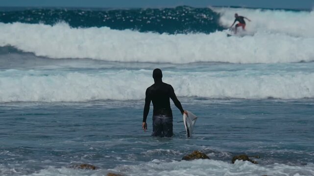 Shirtless Man with surfboard Wading Into Rolling Waves Across Shallow Reef And Rocky Shore, Relaxed Posture With Playful