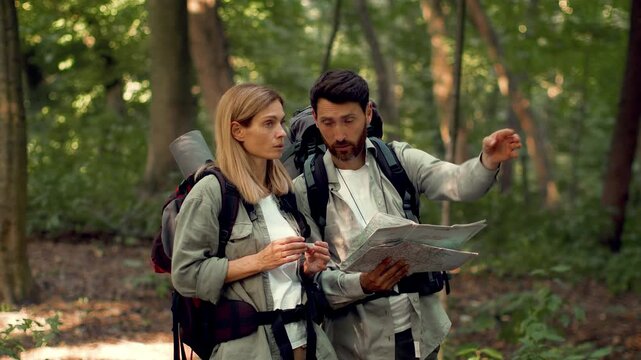 A man and woman look at a map while standing in a forest. They plan their hiking route surrounded by green trees and nature. Their backpacks indicate they are on an outdoor adventure.