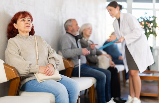 Group of elderly people sitting in chairs in queue to see doctor, nurse talking with patients in background in lobby big medical facility
