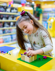 Fototapeta premium Girl concentrating while assembling colorful building bricks green baseplate yellow table toy shop