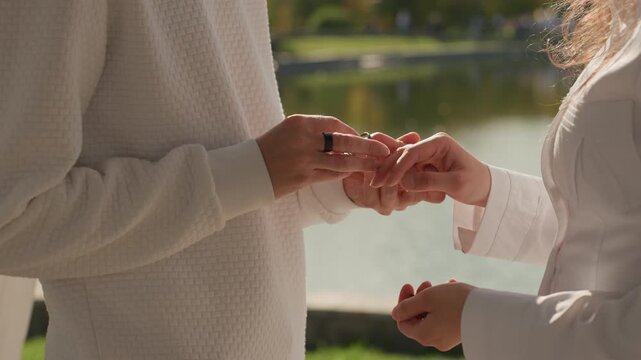 Closeup ring exchange between caucasian partners. Sunlit hands glide a band onto finger near a peaceful waterfront, detailed jewelry shot capturing tenderness, subtle emotion and symbolic promise