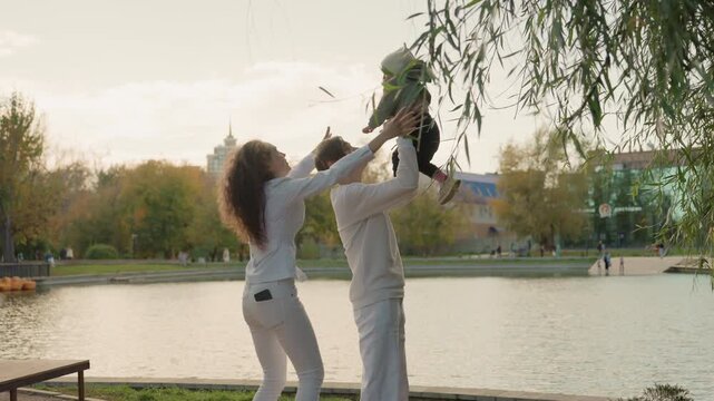 Parents toss baby near shore, playful weekend outing with family and marriage vibe, couple laughing with urban skyline backdrop, bench and riverside trees, candid lifestyle moment