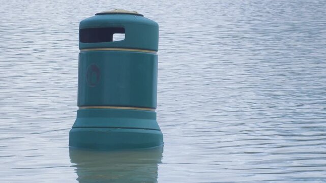 rubbish bin aka trash can surrounded by flood waters, poking up from surface in city park, mute swan swims past in background