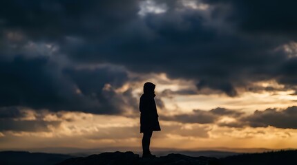Obraz premium Silhouette of person standing against dramatic cloudy sky at sunset with golden light breaking through storm clouds creating moody atmospheric scene.