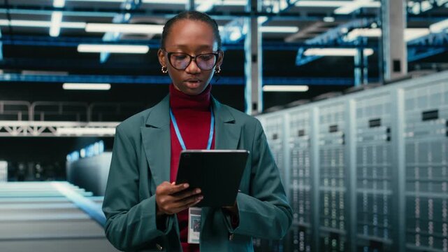 Female engineer walks in data center, using tablet and AR tech to monitor error logs, debugging systems. African american woman in server hub running diagnostics on blue wireframe mesh rigs, camera B