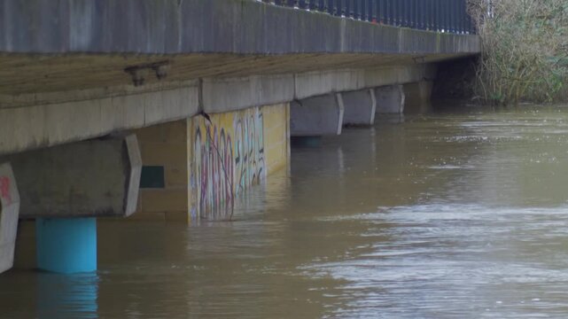 high level of flood water passing through concrete supports of road bridge, riverside city park