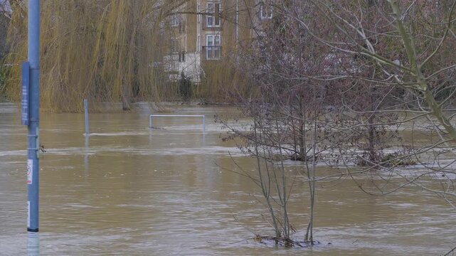 Fast flowing flooded river, muddy brown water, willow trees and residential apartments in background, submerged railing, signpost, lamppost and trees in foreground