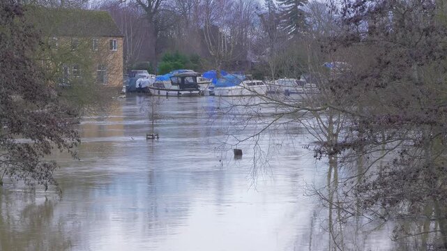 flooded riverside park, boats and building in background, trees surrounded by fast flowing flood waters