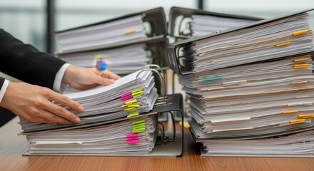 A person in a suit is organizing paperwork in large binders on a desk in an office setting with natu