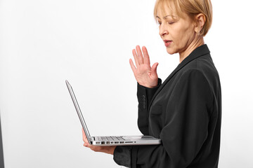 Mature woman in black blazer holds laptop and gestures with hand while participating in video call. Concept for remote work, online communication, and professional meeting.