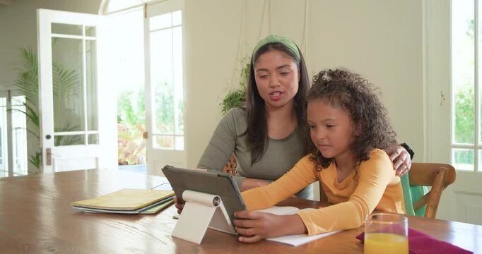 Mother guiding daughter from tablet to paper at home table, pointing while girl practicing writing