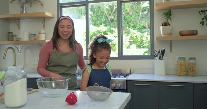 Mother and daughter mother tying girl's apron scooping flour into sifter at kitchen island baking