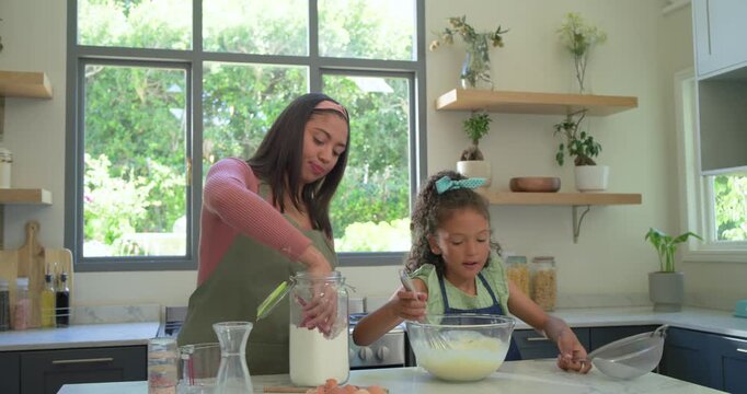 Adult woman and girl in kitchen, woman bringing jar, scooping while girl whisking batter to bake