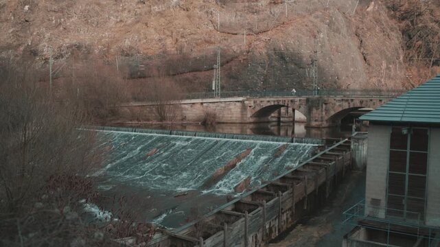 River dam with flowing water and small waterfall near stone bridge and rocky hillside. Industrial hydropower structure in natural landscape under soft daylight, urban infrastructure