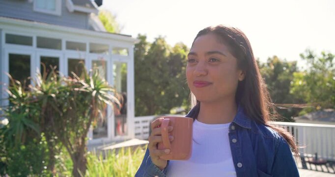 Adult female standing on sunlit deck, lifting pink mug and sipping while relaxing in sunlight