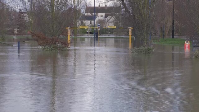 Flooded car park parking lot, flowing flood water, wooded riverside park with trees, houses in distance