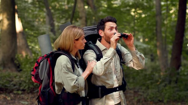 Exciting scene. Emotional man tourist looking forward through binocular and enjoying view, envious woman want to look too, standing together at forest, slow motion