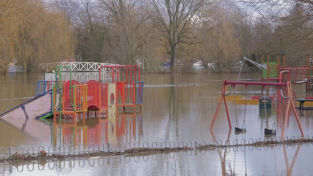 Flooded children's playground, swings, climbing frame, play area submerged in flood water, trees, river and boats in distance