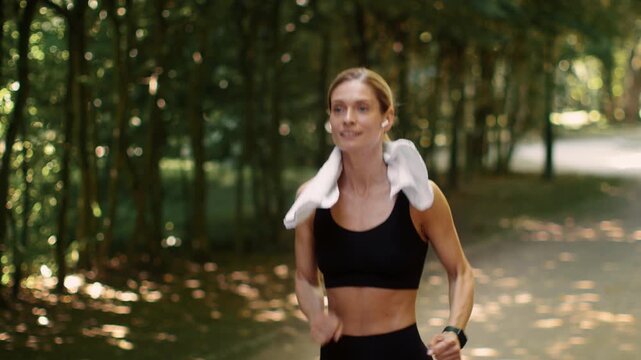 Intensive cardio training. Follow shot of active middle aged lady jogger running in public park and looking at her smartwatch, getting tired and slowing down, slow motion