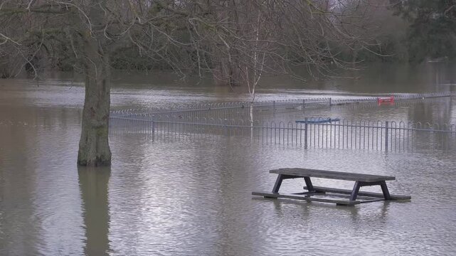 Flooded town riverside park, submerged picnic table, fences, trees, rippling flood water