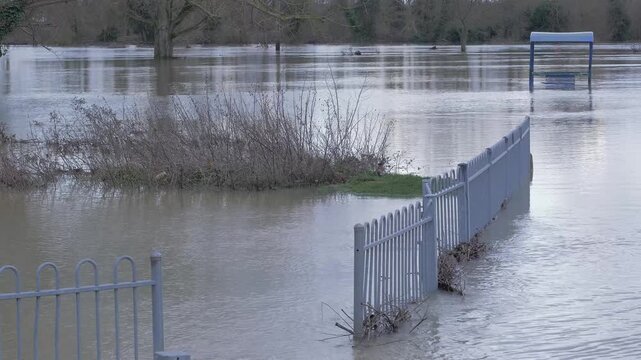 Submerged fence in flooded town park, riverside flood waters, trees and covered bench in background