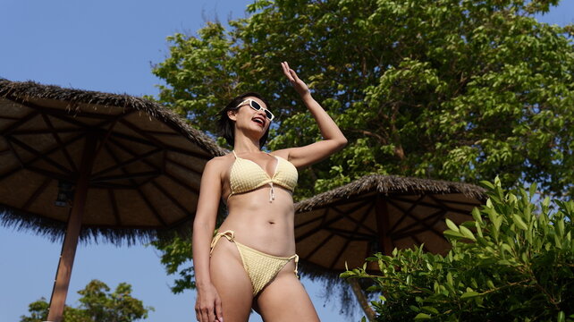 A woman in a yellow bikini and white sunglasses smiles and waves her hand while standing in a garden with thatched palapas