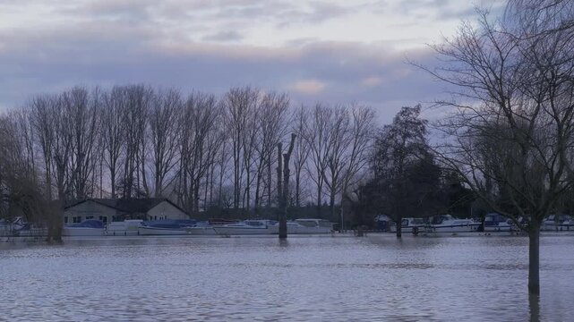 flooded riverside park in later winter light, trees, boats and buildings in background, cloudy stormy sky