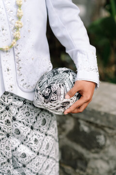 Close up of an Indonesian groom holding a traditional Javanese blangkon headwear, wearing white wedding attire and batik jarik cloth
