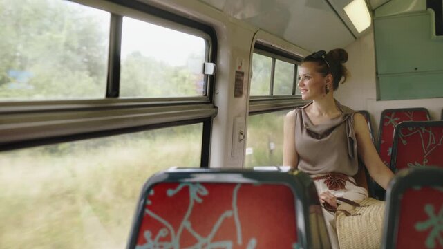 A relaxed young woman sits on a train with red patterned seats, gazing out at the passing green landscape during her summer trip to France.