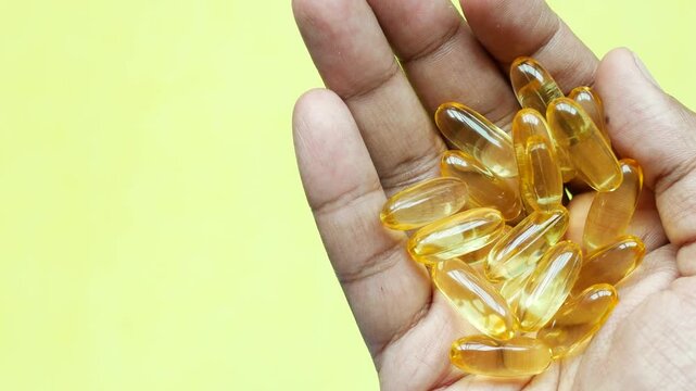 Holding handful of yellow oval vitamin capsules in palm against bright yellow background close up