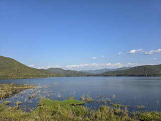 Fototapeta premium Serene landscape view of calm lake and green mountain range under clear blue sky. beautiful and peaceful nature background of water and forest on sunny day