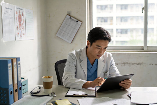 A doctor in a white coat works on a digital tablet at a desk in an office.