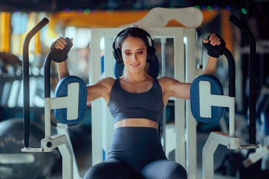 Fit Woman Wearing Headphones Trains on a Pec Deck. Athletic girl with over-ear headphones exercises on chest fly equipment
