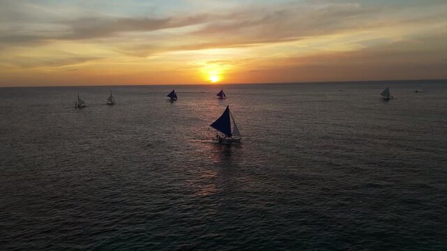 Cinematic aerial shot of traditional paraw sailboats cruising the calm waters of Boracay, Philippines, during a vibrant golden sunset