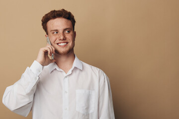 Smiling young man with red hair wearing white shirt talking on smartphone against beige background....